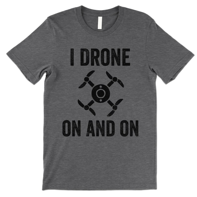 Op de afbeelding: Een donkergrijs T-shirt met de tekst "I DRONE ON AND ON" in zwart. Een zwart drone-icoon is gecentreerd tussen de twee regels tekst.