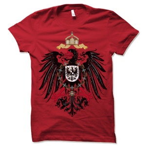 May include: Red t-shirt with a black and gold eagle design. The eagle is holding a shield with a black cross on a white background. The eagle is surrounded by a wreath of gold and red flowers.