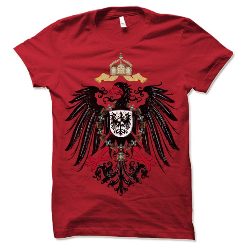 May include: Red t-shirt with a black and gold eagle design. The eagle is holding a shield with a black cross on a white background. The eagle is surrounded by a wreath of gold and red flowers.