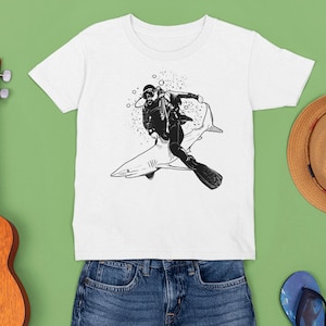 May include: White t-shirt featuring a black and white illustration of a scuba diver riding a shark. The diver is wearing a diving suit and gear. The shirt is displayed with a ukulele, a straw hat, and blue denim shorts.