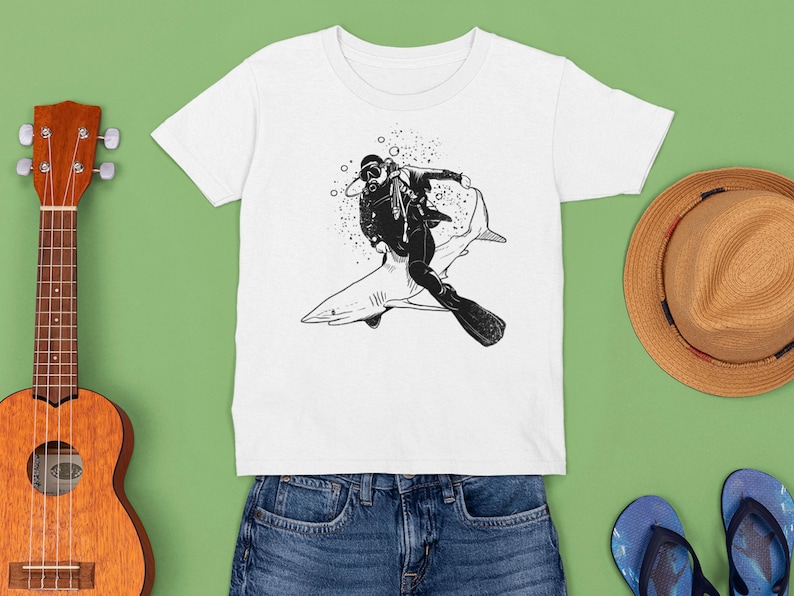 May include: White t-shirt featuring a black and white illustration of a scuba diver riding a shark. The diver is wearing a diving suit and gear. The shirt is displayed with a ukulele, a straw hat, and blue denim shorts.