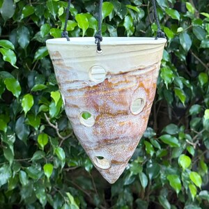 May include: A conical ceramic hanging planter with a cream and brown mottled glaze finish. The planter features several circular cutouts along its sides and is suspended by three thin black cords attached to the upper rim.