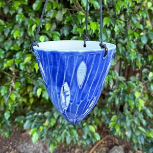 May include: A cone-shaped ceramic hanging planter with a glossy cobalt blue glaze and vertical white textured stripes. The planter features a white interior and is suspended by three thin black cords attached to the rim.
