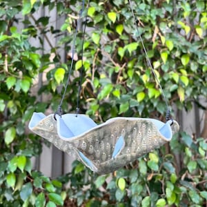 May include: A hanging ceramic bird feeder with a light blue interior and a textured, patterned exterior. The feeder is suspended by black cords against a backdrop of green foliage.