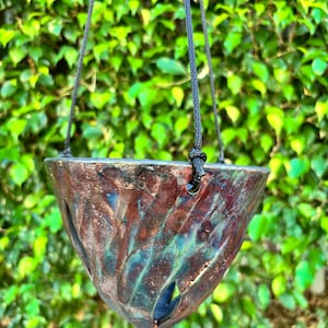 May include: A hanging planter with a triangular shape, featuring a textured surface with shades of brown, red, and teal. The planter is suspended by black rope, with a backdrop of green foliage.