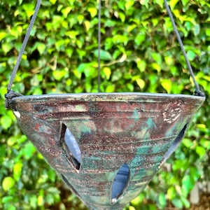May include: A hanging planter with a triangular shape, featuring a blend of brown, green, and red hues. The planter has several oval-shaped openings and is suspended by a black rope. The background is a dense wall of green foliage.