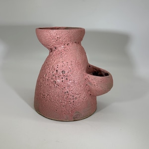 Op de afbeelding: Een roze keramische vaas met een getextureerd, putjesoppervlak. De vaas heeft een bolvormige basis, een smalle hals en een kleine, afgeronde zijopening. De kleur is een gedempt, stoffig roze, en de algehele vorm is organisch en sculpturaal.