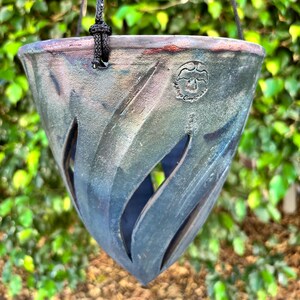 May include: Hanging ceramic planter with a dark, textured finish and a unique, carved design. The planter has a flared shape and is suspended by a black rope. The background is a lush green wall of leaves.