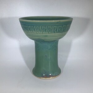 Può includere: Una ciotola in ceramica artigianale, in stile piedistallo, nei toni del verde e del blu. La ciotola ha un'ampia parte superiore aperta e una fascia strutturata al centro. La base è di un verde più chiaro a contrasto, con un piede leggermente svasato. La ciotola è alta circa 20 cm.