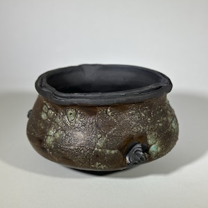 Op de afbeelding: Een kleine, ronde keramische kom met een getextureerde, gevlekte bruine buitenkant en een zwart interieur. De kom heeft een donkere rand en twee kleine, decoratieve handvatten. De buitenkant heeft groene en blauwe vlekken.