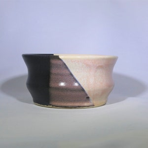 May include: A small ceramic bowl with a black, pink, and white glaze. The bowl has a rounded shape and a slightly flared rim.