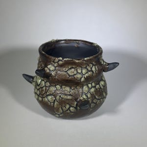 May include: A handcrafted ceramic vessel with a textured, cracked surface in shades of brown and cream. The small pot features two dark gray, horn-like protrusions and a dark interior. The handmade pottery is suitable for home decor.