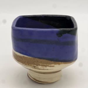 May include: A small, square ceramic bowl with a wide, flat base. The bowl is decorated with a blue glaze and black stripes, with a brown glaze around the base.