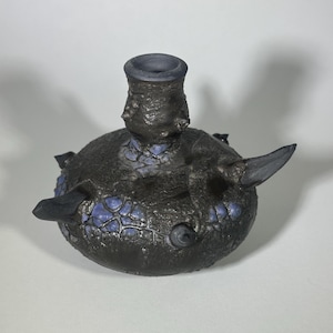 May include: A handcrafted ceramic vessel with a dark, textured exterior and a cylindrical neck. The spherical body features blue accents and wing-like protrusions. The vessel is likely used for decorative purposes.