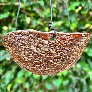 May include: A brown ceramic hanging planter with a textured, pebble-like surface. The planter is suspended by black wire and has a hole for drainage. The background is blurred green foliage, suggesting an outdoor setting. Ideal for small plants.
