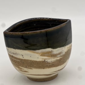 May include: A small, ceramic cup with a dark green and brown glaze. The cup has a white band with brown streaks around the middle.