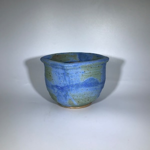 May include: A small, blue and green ceramic planter with a hexagonal shape. The planter has a speckled glaze and a slightly uneven rim.
