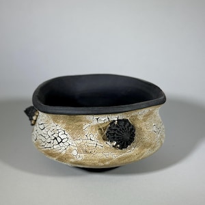 Puede incluir: Un cuenco de cerámica hecho a mano con interior negro y exterior texturizado blanquecino. El exterior presenta un esmalte craquelado y dos adornos decorativos negros. Una pieza de cerámica única.