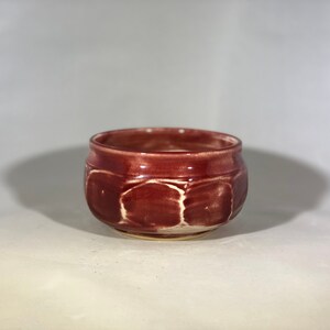 May include: A small, red ceramic bowl with a textured, slightly raised design. The bowl has a rounded shape and a slightly flared rim.