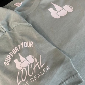 Puede incluir: Dos camisetas verde claro con gráficos blancos. Una camiseta tiene un diseño de cerámica y una taza. La otra camiseta tiene el texto "SUPPORT YOUR LOCAL POT DEALER" con el mismo gráfico de cerámica. Las camisetas son de un material suave.