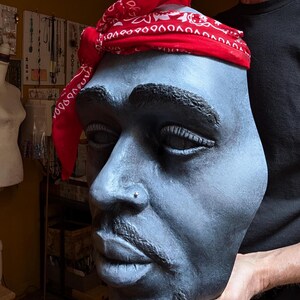 May include: A sculpted bust of a man with a red bandana. The bust is painted in shades of gray and has a detailed depiction of facial features, including a nose ring and facial hair. The red bandana is tied around the head.