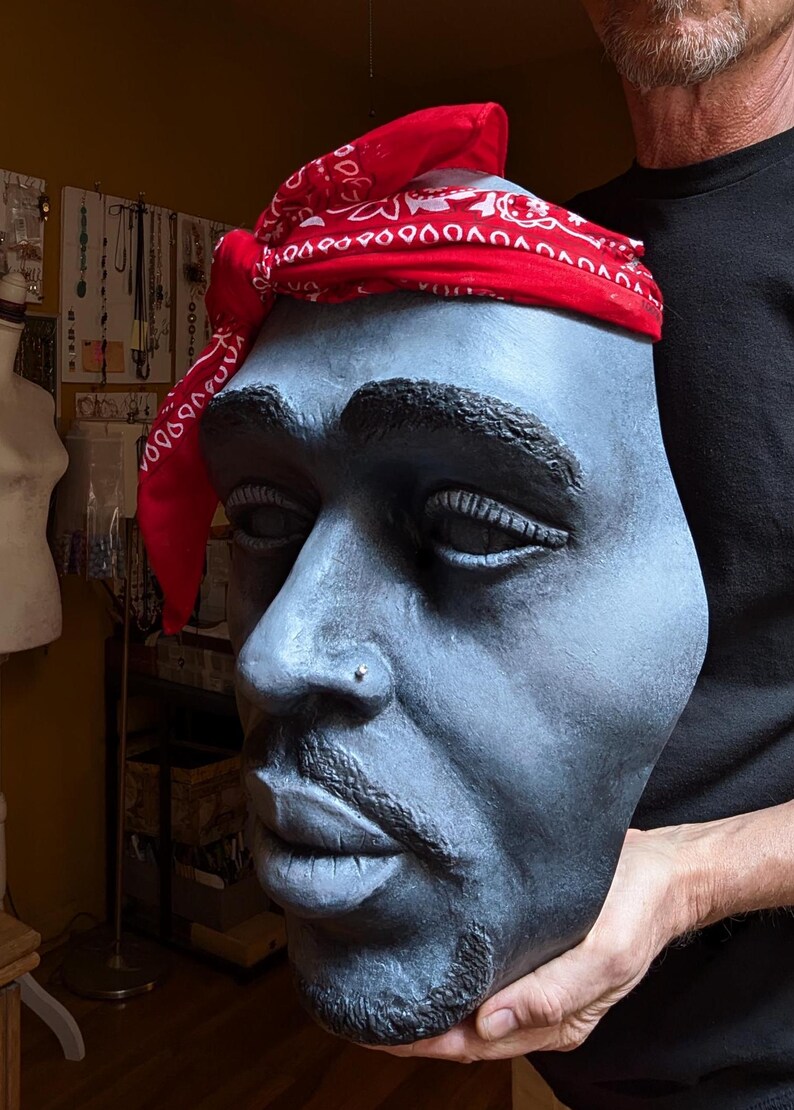 May include: A sculpted bust of a man with a red bandana. The bust is painted in shades of gray and has a detailed depiction of facial features, including a nose ring and facial hair. The red bandana is tied around the head.