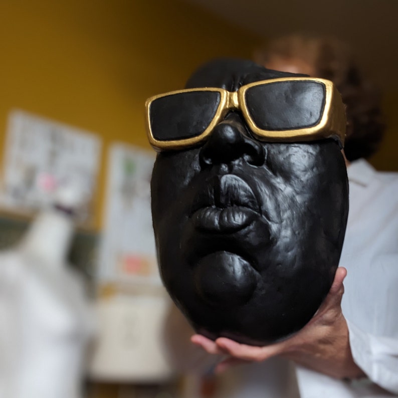 May include: A black sculpture of a face wearing gold-rimmed sunglasses. The sculpture has a detailed facial structure with prominent lips and a rounded shape. The background is a blurred yellow wall.