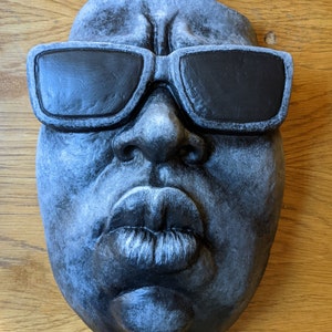 May include: A gray, sculpted face with black sunglasses. The face has full lips and a detailed nose. The sculpture is likely made of stone or a similar material, and the sunglasses are painted black. The overall aesthetic is artistic and expressive.