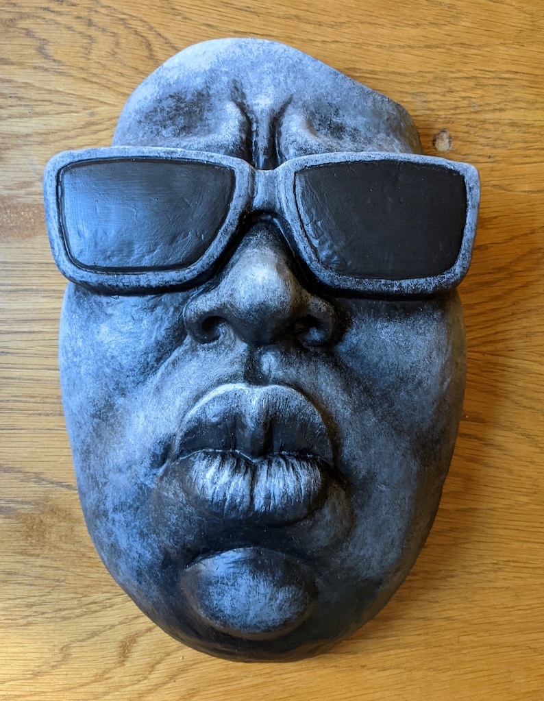 May include: A gray, sculpted face with black sunglasses. The face has full lips and a detailed nose. The sculpture is likely made of stone or a similar material, and the sunglasses are painted black. The overall aesthetic is artistic and expressive.