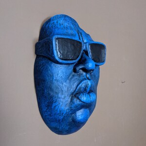 May include: A blue wall sculpture of a face wearing black sunglasses. The sculpture has a textured surface and is mounted on a neutral-colored wall. The face has prominent features, including a large nose and full lips.