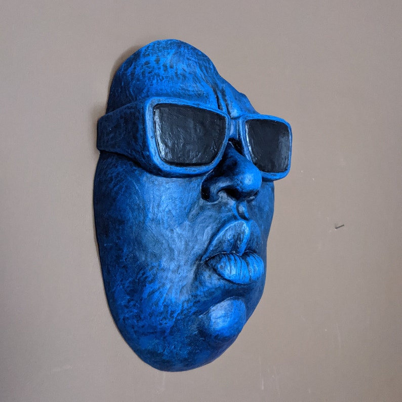 May include: A blue wall sculpture of a face wearing black sunglasses. The sculpture has a textured surface and is mounted on a neutral-colored wall. The face has prominent features, including a large nose and full lips.