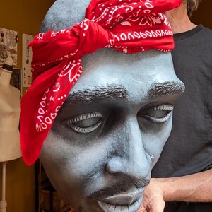 May include: A sculpted bust of a man with closed eyes, painted in shades of gray and black, wearing a red bandana with a white paisley pattern. The bust is held by a person, showcasing the detailed facial features.