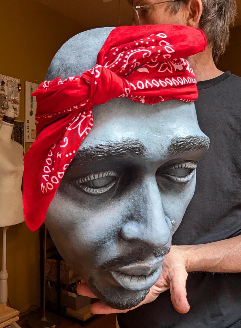 May include: A sculpted bust of a man with closed eyes, painted in shades of gray and black, wearing a red bandana with a white paisley pattern. The bust is held by a person, showcasing the detailed facial features.