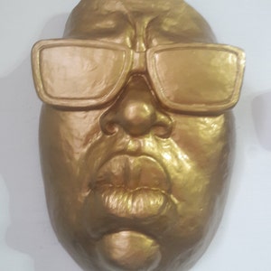 May include: A golden-colored mask with sunglasses. The mask has a detailed face with a prominent nose, full lips, and square-framed sunglasses. The mask is likely a decorative item or a piece of art.