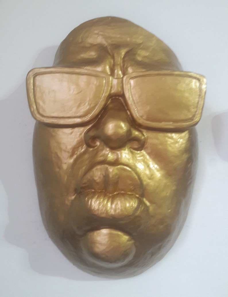 May include: A golden-colored mask with sunglasses. The mask has a detailed face with a prominent nose, full lips, and square-framed sunglasses. The mask is likely a decorative item or a piece of art.