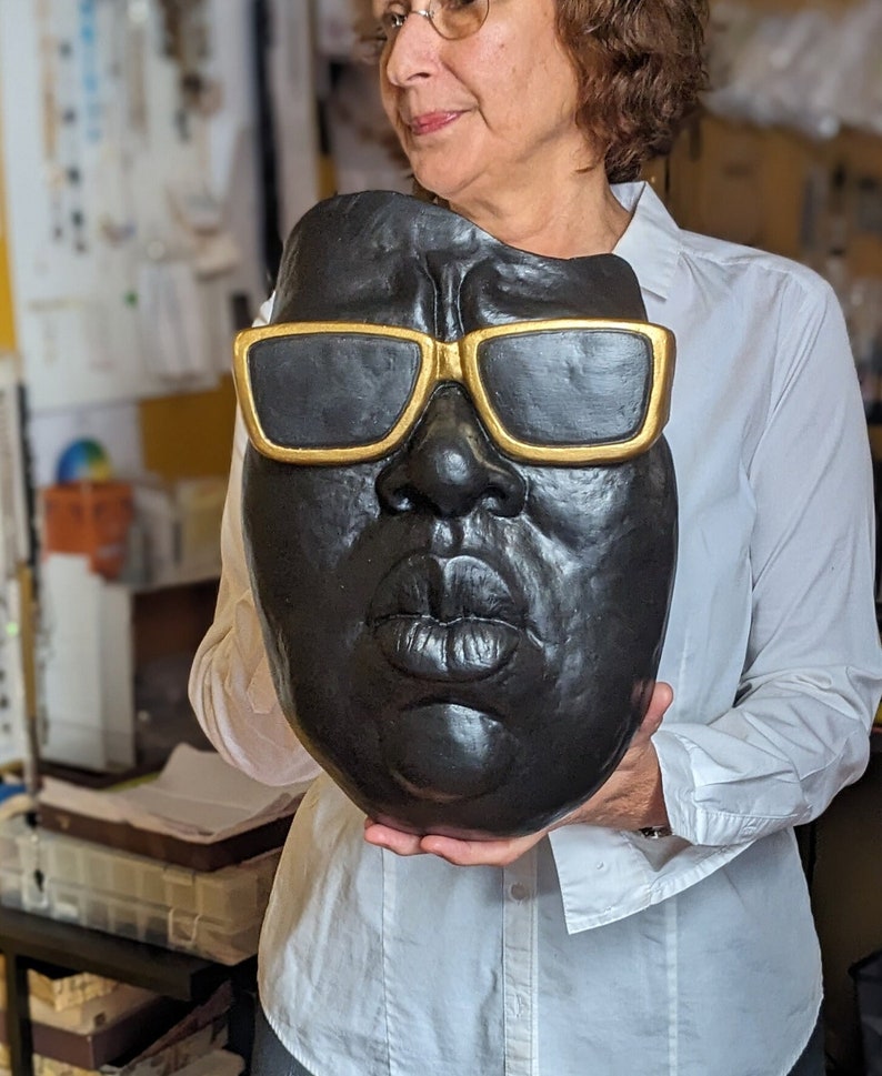 May include: A black sculpture of a face with gold-rimmed sunglasses. The sculpture has a textured surface and is held by a person wearing a white shirt. The sculpture's features include a prominent nose and full lips.