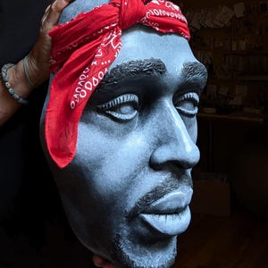 May include: A sculpted bust of a man with a red bandana tied around his head. The bust is painted in shades of gray and has a goatee. The bandana has a white pattern. The bust is held by a person.