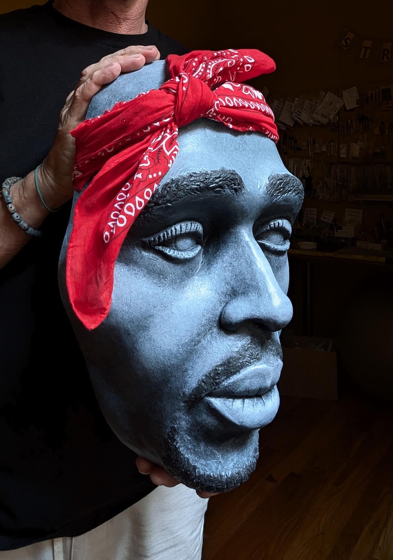 May include: A sculpted bust of a man with a red bandana tied around his head. The bust is painted in shades of gray and has a goatee. The bandana has a white pattern. The bust is held by a person.