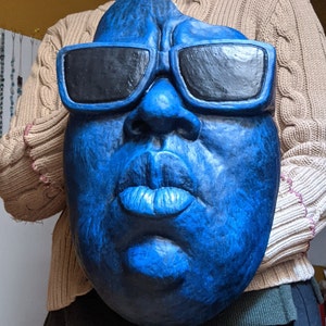May include: A large, blue face sculpture with black sunglasses. The sculpture has a textured surface and detailed facial features, including a prominent nose and full lips. The sunglasses are rectangular and block the eyes. The sculpture is held by a person.