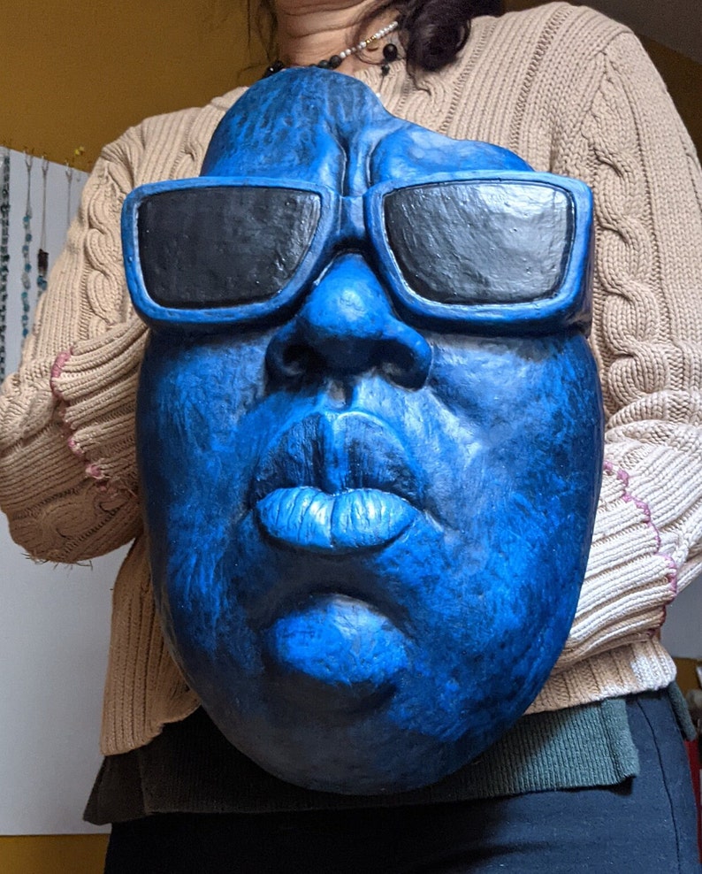 May include: A large, blue face sculpture with black sunglasses. The sculpture has a textured surface and detailed facial features, including a prominent nose and full lips. The sunglasses are rectangular and block the eyes. The sculpture is held by a person.