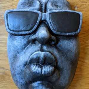 May include: A gray, sculpted face with black sunglasses. The face has full lips and a textured surface. The sculpture is mounted on a wooden surface.
