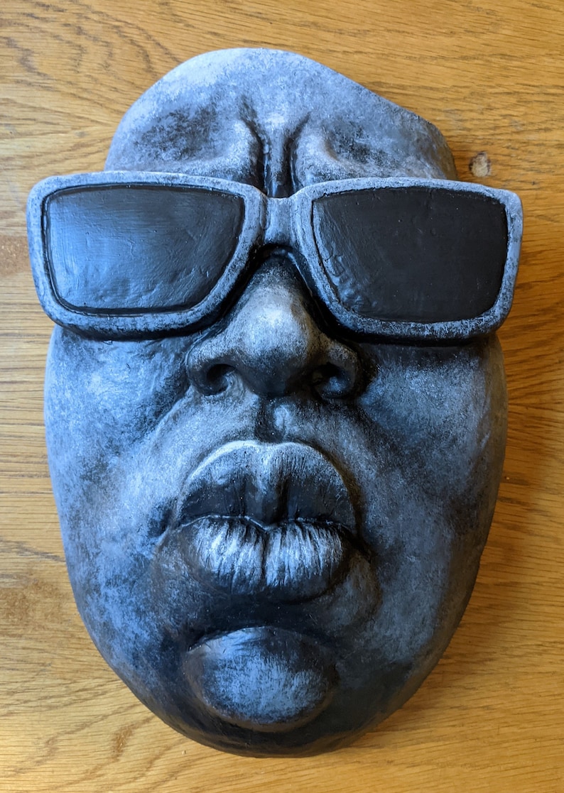May include: A gray, sculpted face with black sunglasses. The face has full lips and a textured surface. The sculpture is mounted on a wooden surface.