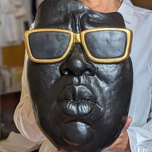 May include: A black mask with gold-rimmed sunglasses. The mask features a detailed depiction of a face with full lips and a prominent nose. The mask is held by a person wearing a white shirt.