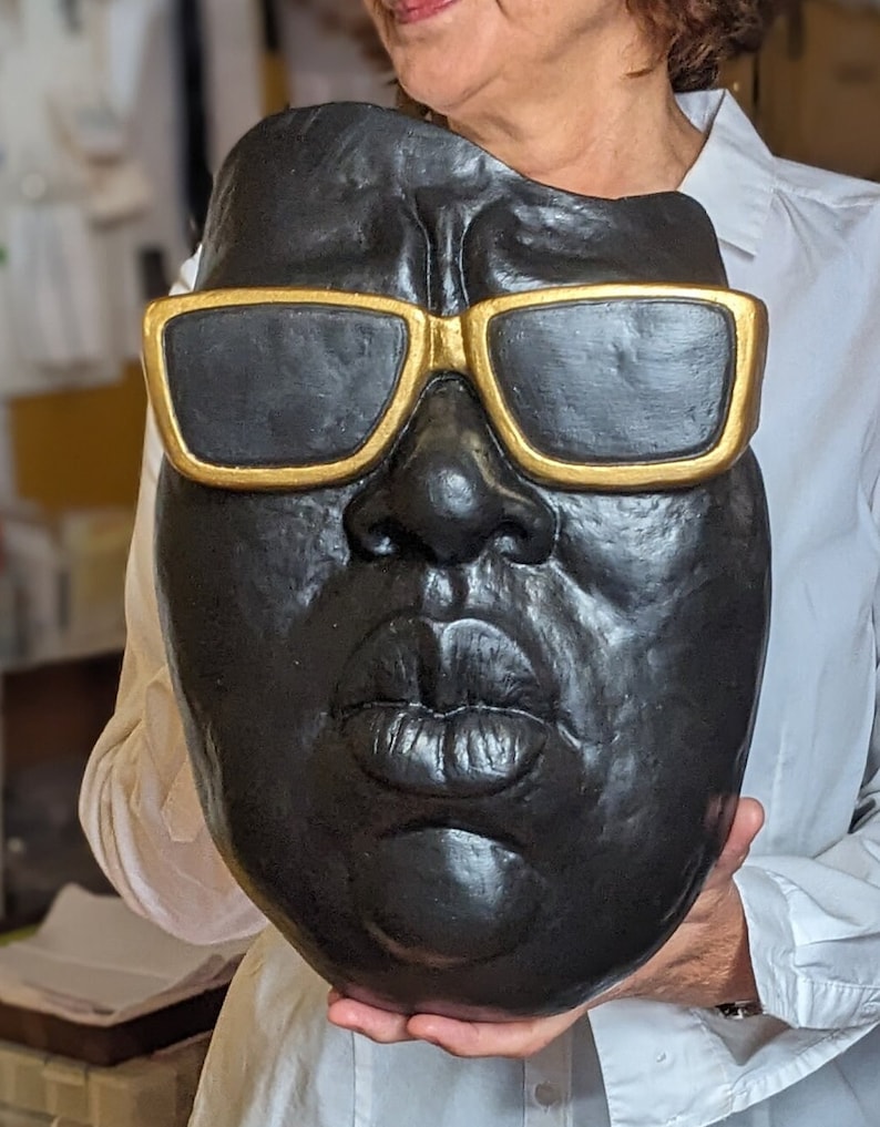 May include: A black mask with gold-rimmed sunglasses. The mask features a detailed depiction of a face with full lips and a prominent nose. The mask is held by a person wearing a white shirt.