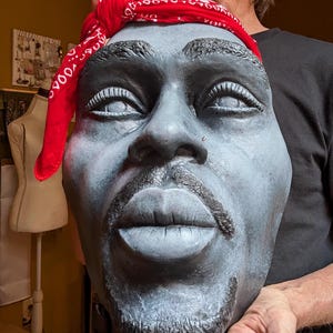 May include: A gray sculpture of a face with a red bandana. The sculpture has a goatee and mustache. The bandana is red with a white pattern. The sculpture is held by a person.