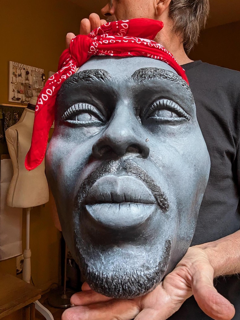 May include: A gray sculpture of a face with a red bandana. The sculpture has a goatee and mustache. The bandana is red with a white pattern. The sculpture is held by a person.
