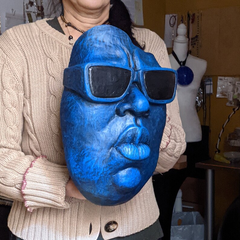 May include: A large, blue sculpture of a face wearing black sunglasses. The face has prominent lips and a textured surface. The sculpture is held by a person wearing a beige sweater.