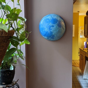 Könnte beinhalten: Ein rundes, blaues Wandkunstwerk, das einem Planeten ähnelt und an einer mauvefarbenen Wand befestigt ist. Das Kunstwerk weist strukturierte Details und verschiedene Blautöne auf. Eine Topfpflanze mit grünen Blättern ist ebenfalls zu sehen.