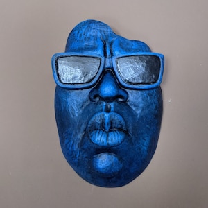 May include: A blue sculpted face with black sunglasses. The face has a prominent nose and full lips. The sculpture is a solid blue color and appears to be made of a textured material.