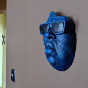 May include: A blue wall sculpture of a face wearing black sunglasses. The sculpture is mounted on a neutral-colored wall. The face has a prominent nose and lips. The sculpture is a unique piece of wall art.